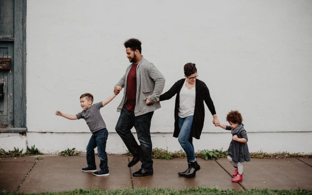 Photo by Emma Bauso via Pexels. A family of four walk down a sidewalk holding hands. A little boy leads the way, dragging his father, who’s wearing a gray sweater and a read shirt, forward. A smaller girl in a gray dress and red boots walks at the end of their line, holding her mother’s hand as she looks back to check on her.